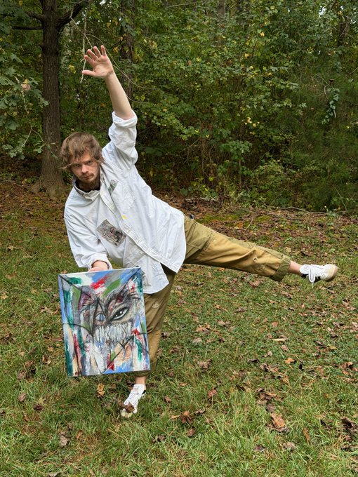 A young man with curly hair posing dramatically outdoors in a white jacket and olive pants, balancing on one leg while holding a colorful abstract painting of a face in a grassy, wooded area.