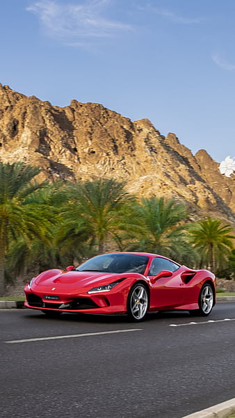 A red Ferrari sports car parked on a road with rugged mountains and palm trees in the background under a blue sky.