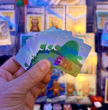 A hand fans out a set of collectible holographic cards featuring Care Bears artwork, with a display of Bearbrick figurines visible in the blurred background.
