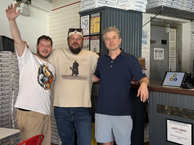 Three men pose cheerfully at Williamsburg Pizza, where stacks of branded pizza boxes line the walls, connecting to the "Doge & Pizza" post with their casual, fun pizza-parlor vibe.