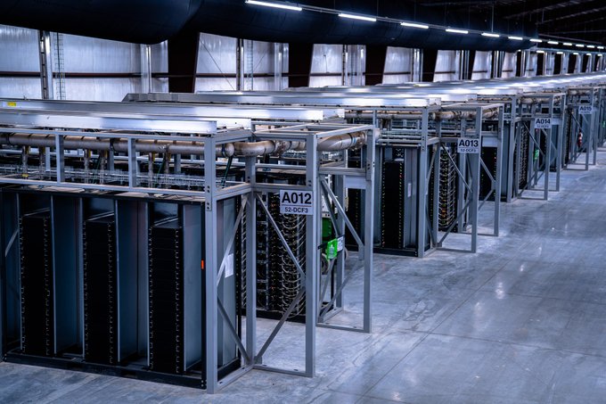 Rows of empty server racks labeled A012, A013, and A014 stand illuminated inside a large, industrial data center facility.