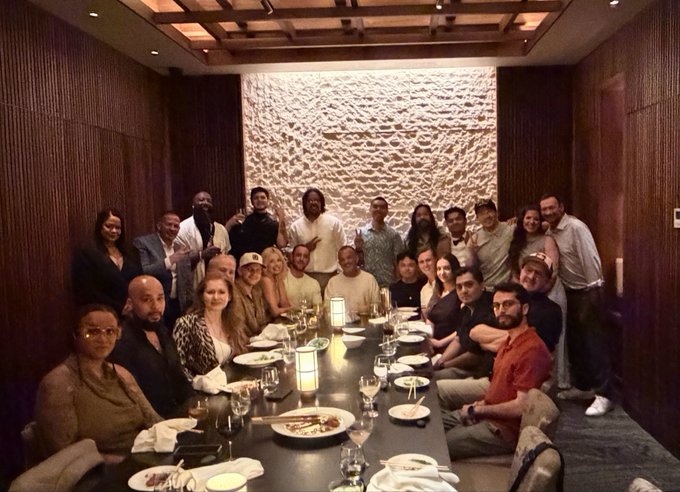 A large group of people gathered for a group photo around a long dining table in an upscale restaurant with a textured white wall feature.