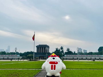 A white inflatable mascot character with a red bull logo stands on a grassy field in front of what appears to be the Ho Chi Minh Mausoleum in Hanoi, Vietnam, with the Vietnamese national flag visible. The image illustrates the opening of a travel series documenting a journey through Vietnam's iconic landmarks, beginning at this historic site.