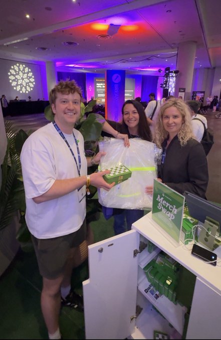 The image shows three people standing next to a white cabinet, each holding an item that appears to be merchandise from Matcha.xyz. The person on the left is holding a green box with a white label, while the other two individuals are holding items in their hands. They seem to be posing for a photo or attending an event related to Matcha products.