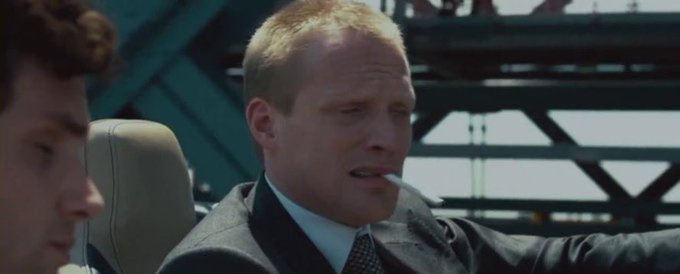 A cinematic still of a man with a shaved head smoking a cigarette in a car, with another passenger partially visible beside him.