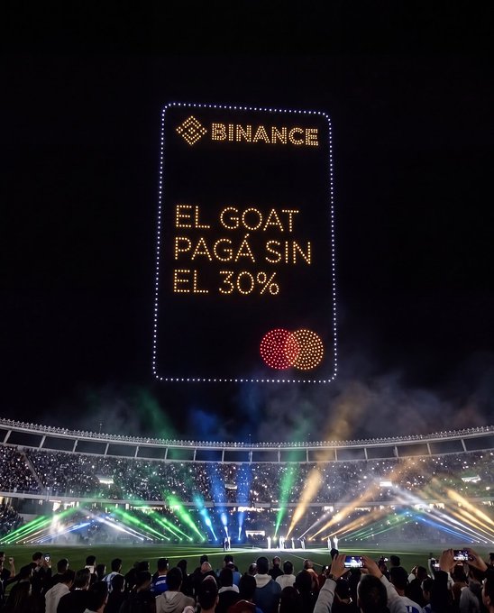A stadium screen displays a Binance credit card-shaped advertisement in lights reading 'EL GOAT PAGA SIN EL 30%' during a colorful light show.