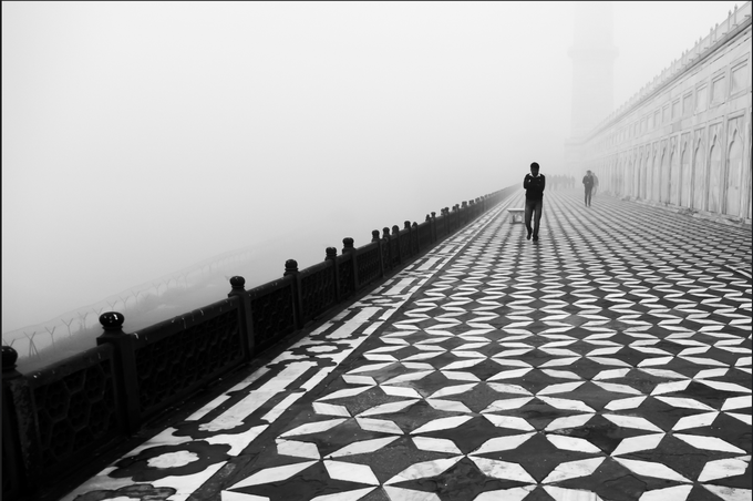A black and white photograph of a foggy walkway with a geometric star-patterned floor, where a solitary figure walks away into the mist.