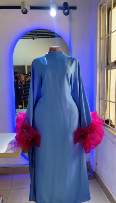 A mannequin displays a long-sleeved dusty blue gown with bright pink ruffled cuffs, photographed in front of an oval mirror with blue LED lighting in a shop setting.