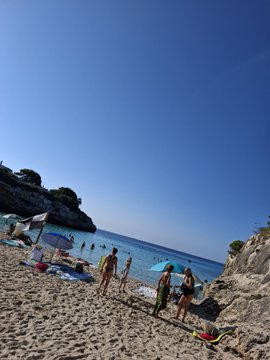 A sunny beach scene with people walking on sand and swimming in clear blue water under a bright blue sky.