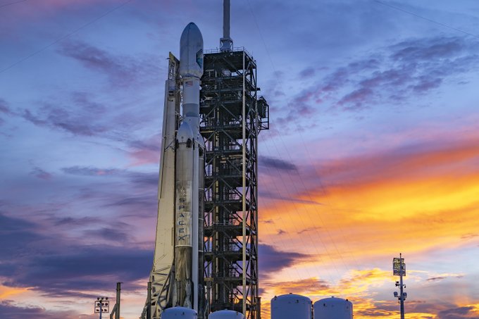 A SpaceX Falcon 9 rocket with the Crew Dragon spacecraft standing on its launch pad against a dramatic sunset sky with purple, orange, and blue clouds.