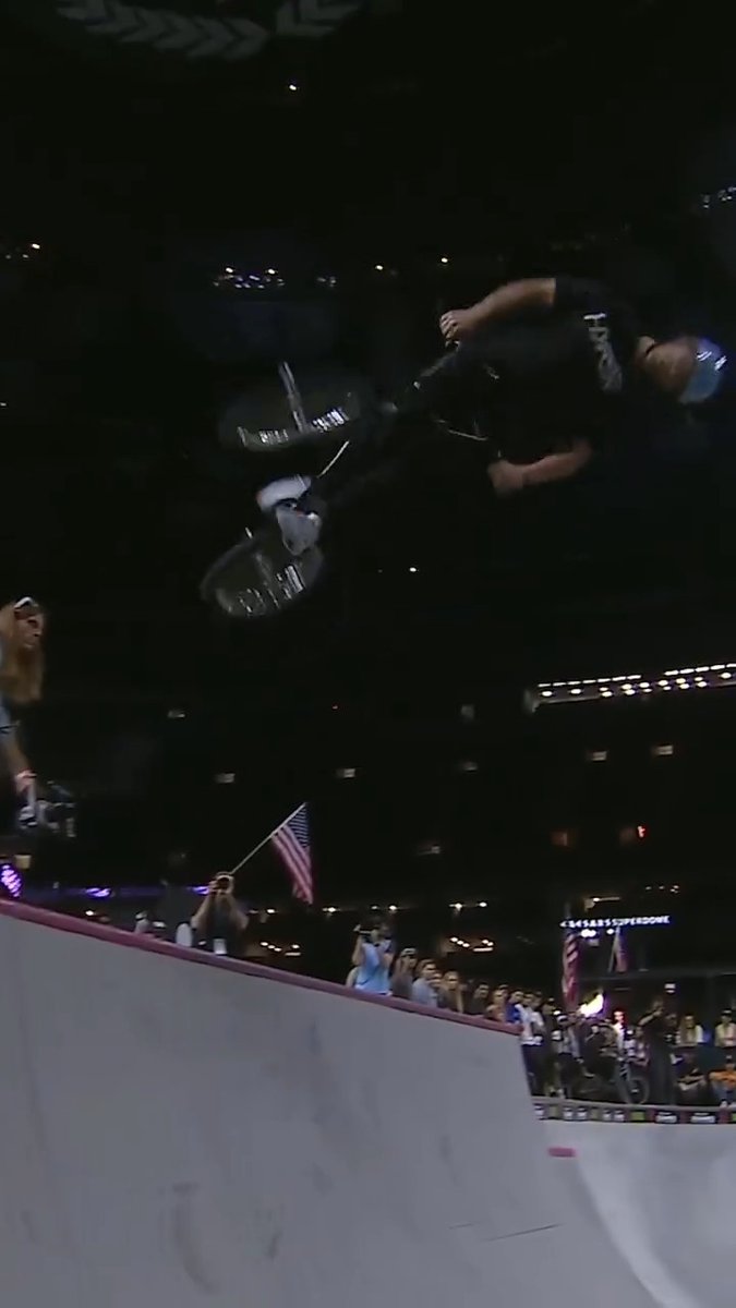 A nighttime BMX rider performing a high aerial trick above a halfpipe ramp at an indoor arena, with spectators and an American flag visible below.