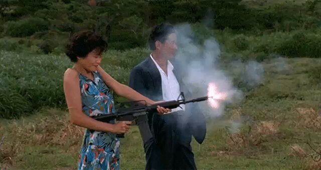 The image shows a woman wearing a blue floral dress holding a gun, while a man in a suit stands next to her with his hands behind his back. The scene appears to be set in an open field or grassy area, possibly during a military exercise or training session.