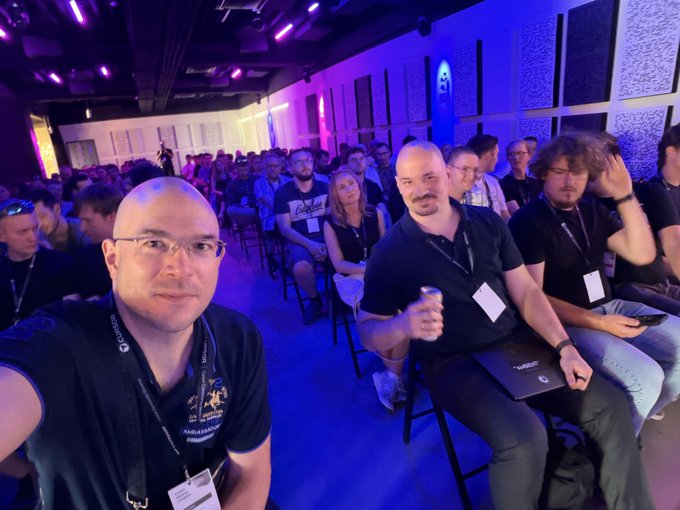 The image shows a group of people sitting in chairs, possibly attending an event or gathering. There are at least 12 people visible in the scene, with some individuals wearing name tags indicating they might be part of a conference or meeting. The room appears to have a blue and white color scheme, creating a calm atmosphere for the attendees.

In addition to the people and chairs, there is an open laptop placed on one of the chairs, suggesting that someone may be using it during the event. A backpack can also be seen in the scene, likely belonging to one of the attendees. The image captures a moment where two men are taking a selfie together with their name tags visible, possibly as part of a group photo or social media post.