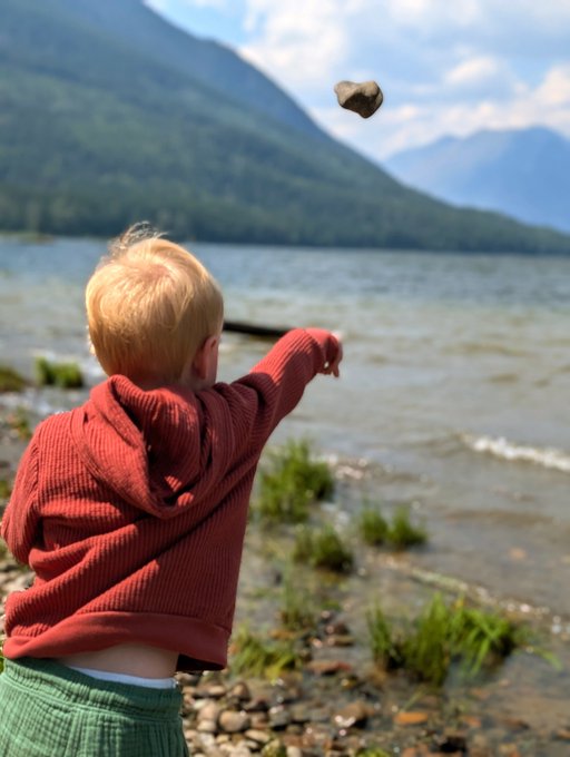 A young child with blonde hair wearing a red hooded sweater and green pants, skipping a rock across a lake with mountains in the background.