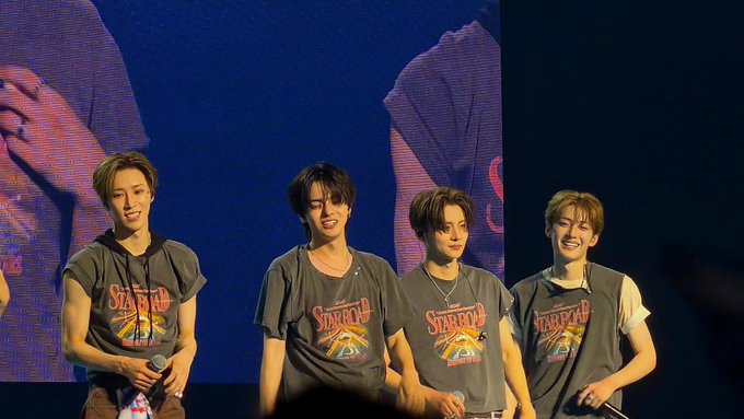 Four members of Stray Kids performing on stage, all wearing matching gray Starbucx graphic t-shirts against a blue-lit backdrop.