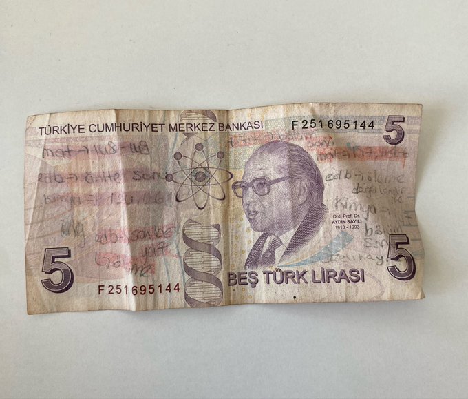 A well-worn, creased 5 Turkish Lira banknote featuring a portrait of Aydın Sayılı, covered in handwritten calculations and notes in red and black ink.