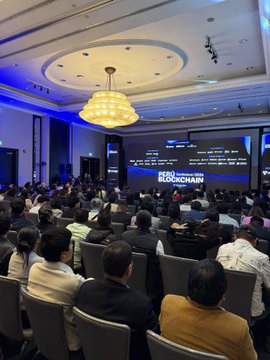 Indoor conference or event venue with attendees seated facing a large screen displaying 'BLOCKCHAIN' text, featuring an ornate circular ceiling with chandelier and blue accent lighting.