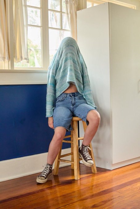 A person sitting on a wooden stool with a striped towel draped over their head to obscure their face, wearing denim shorts, a striped sweater, and black high-top sneakers in a room with blue walls.