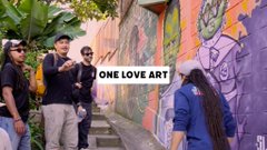 A group of young people posing together outdoors, with one wearing a jacket that displays the text 'ONE LOVE ART'.