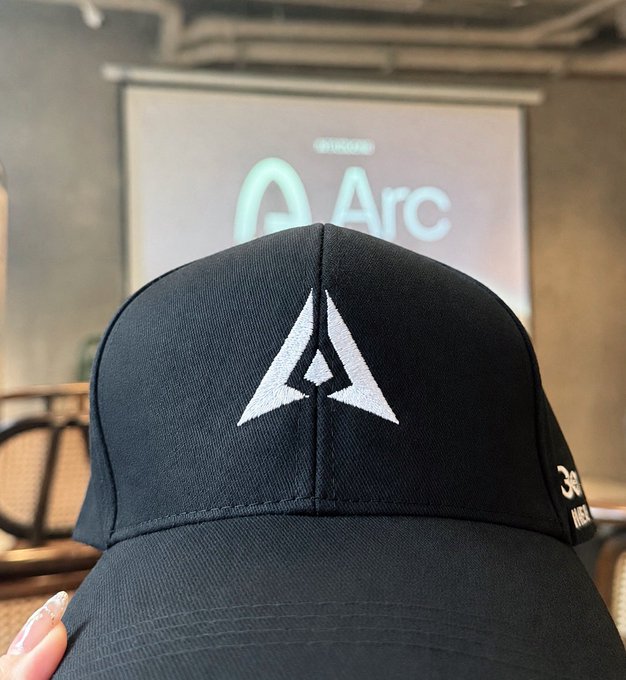 A close-up of a black baseball cap featuring an embroidered triangular logo, with a hand displaying manicured nails visible at the bottom; an 'Arc' branded screen is visible in the background.