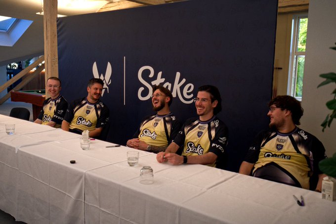 Five players in matching yellow and black "Stake" jerseys laugh together while seated at a long table beneath a backdrop featuring the team's logo and "Stake" branding, apparently showcasing the sponsor's new kit during a media event.