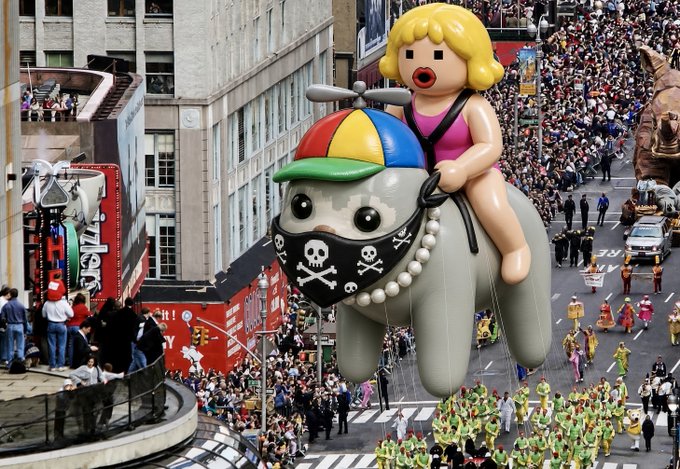 An aerial view of a crowded parade, likely the Macy's Thanksgiving Day Parade, featuring a large balloon of a blonde girl with red lips riding a gray elephant with a skull-and-crossbones bandana.