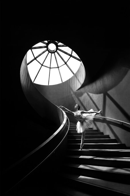 A black and white photograph of a ballerina in a white tutu posed gracefully on a spiral staircase beneath an illuminated circular skylight.