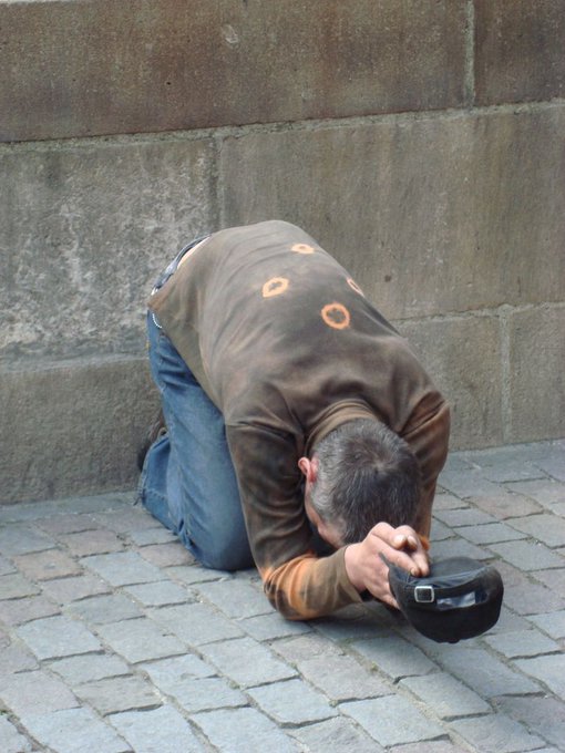 A man wearing a brown patterned shirt and jeans kneeling on a cobblestone street, bowing down with his head to the ground while holding a black hat.