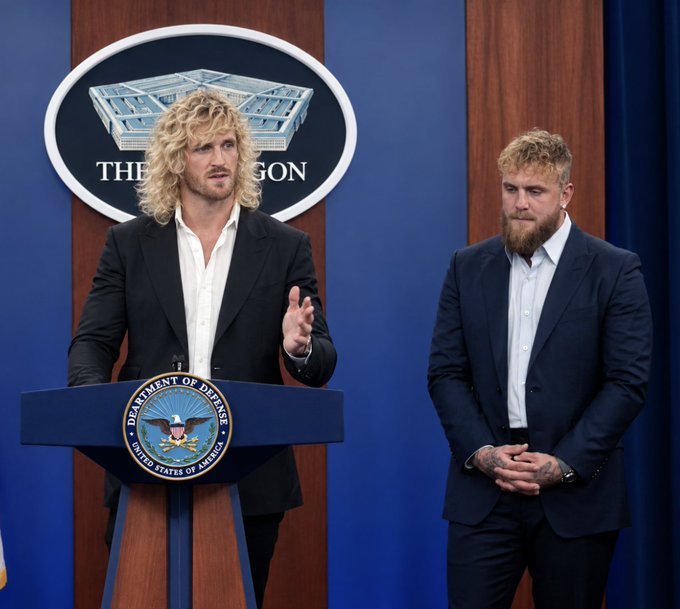 Two men stand at a podium bearing the "Department of Defense United States of America" seal during a Pentagon press briefing. The setup reflects the social media context of Logan Paul (left, speaking) and Jake Paul (right) visiting the U.S. Department of Defense headquarters.