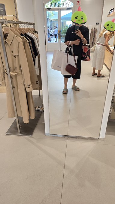Mirror selfie in a Burberry boutique showing a person in black holding shopping bags, their face obscured by a green cactus sticker.