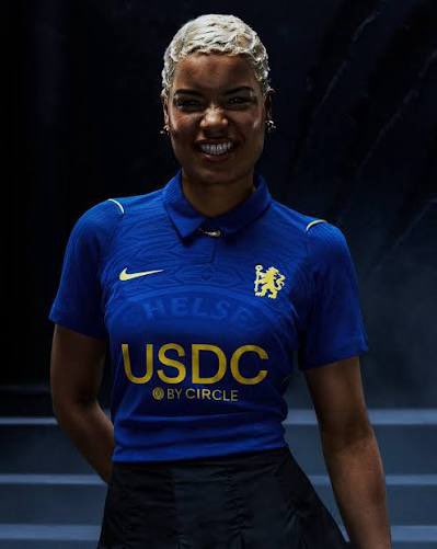 A smiling woman with short blonde hair models a blue Chelsea FC soccer jersey featuring "USDC BY CIRCLE" as the main sponsor on the front.