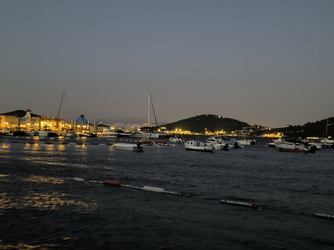 An evening harbor scene with numerous small boats and sailboats anchored in calm water, with illuminated buildings and hills in the background.