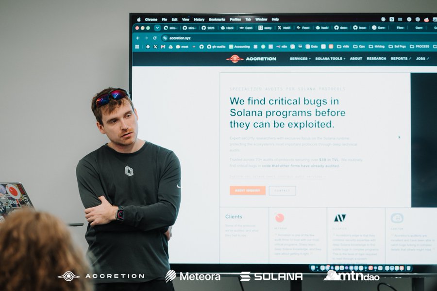 A man stands with crossed arms in front of a large screen displaying the Accretion website, which advertises specialized audits for Solana protocols to find critical bugs.