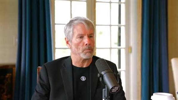 A man with gray hair and a beard, wearing a black jacket over a black shirt with a small logo, is speaking into a microphone during what appears to be a podcast recording.