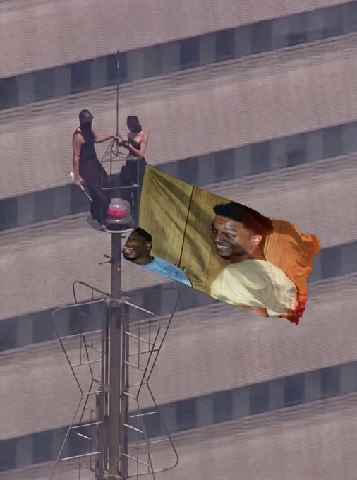 Two people stand on a small aerial platform attached to a broadcast tower, working on a large digital billboard displaying a smiling person's face, set against the facade of a modern building—visually echoing the optimistic "Solana looks good here" sentiment.
