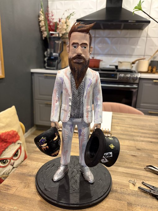 A painted figurine of a bearded man in a light purple suit holding two hats, displayed on a round black base on a wooden table in a kitchen setting.