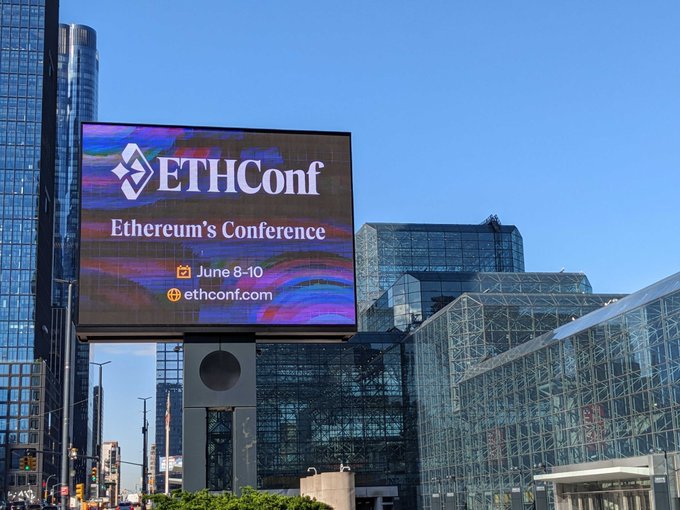 The image shows a large billboard for an upcoming EthCon conference held at the Javits Center in New York City. The billboard features a blue background with white text that reads "ETHCON" and "It's official... 7 days until ETHConf See you at Javits, June 8-10." The backdrop of the billboard is filled with tall buildings, indicating that the event will be held in an urban setting.