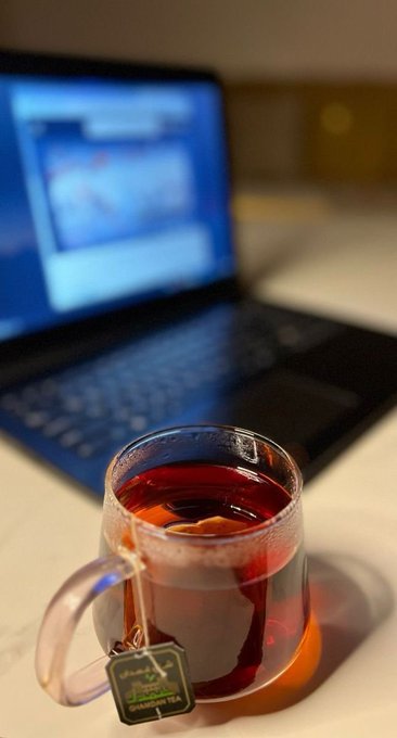 A clear glass mug filled with red tea and a steeping teabag (with a label visible) sits in the foreground, with a blurred laptop displaying a webpage in the background.