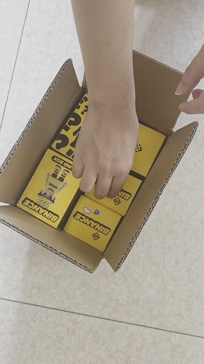 The image shows a person's hand holding a cardboard box filled with toys, specifically action figures. The person appears to be opening or rearranging the items inside the box. The text "尊敬币安美股交易员们" is present in the image as well.
