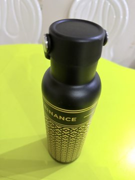 A black insulated bottle with gold patterned design and partial text reading 'NANCE' on a bright green table.