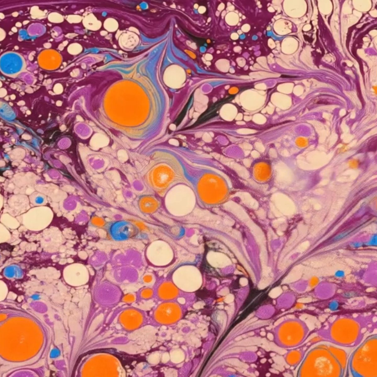 The image features a close-up view of an abstract painting with purple, orange, and white colors. The painting has a unique texture that resembles a marble effect or a marbled pattern. The artist's signature can be seen at the bottom right corner of the image. The background is blurred, drawing attention to the vibrant colors and intricate details in the foreground.