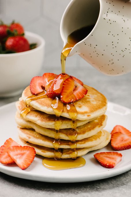 A stack of pancakes topped with fresh strawberry slices is being drizzled with syrup from a white speckled pitcher, with a bowl of whole strawberries visible in the background.