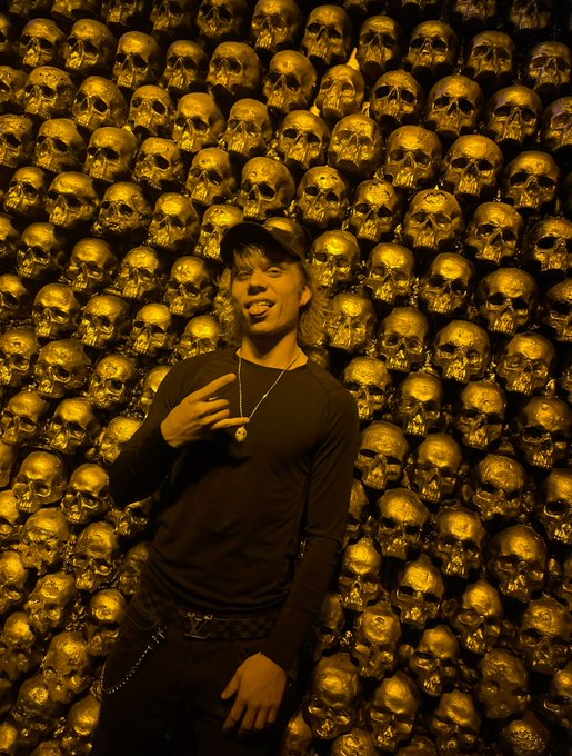 A man wearing a black cap, black long-sleeved shirt, and a pendant necklace poses with his tongue out against a wall entirely covered in gold-colored sculpted skulls, illuminated by warm lighting.