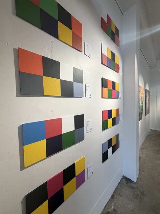 A photograph of a white gallery wall displaying multiple horizontal canvases with colorful geometric grid patterns in primary colors, accompanied by small white information cards next to each piece.