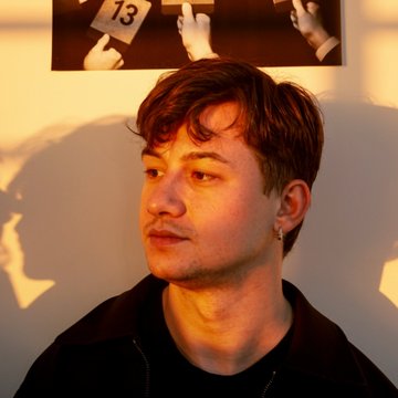 A portrait of a young man with brown hair and a mustache, lit with warm light, with a photograph partially visible on the wall behind him.