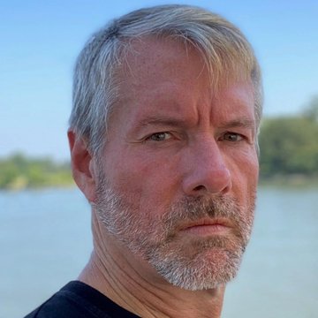 The image features a man with a beard, looking at the camera while standing near water. The background includes trees and a body of water, creating an outdoor setting for this portrait. There are no texts or other objects present in the image, making it a simple yet visually appealing composition that highlights the subject's facial expression and the surrounding environment.