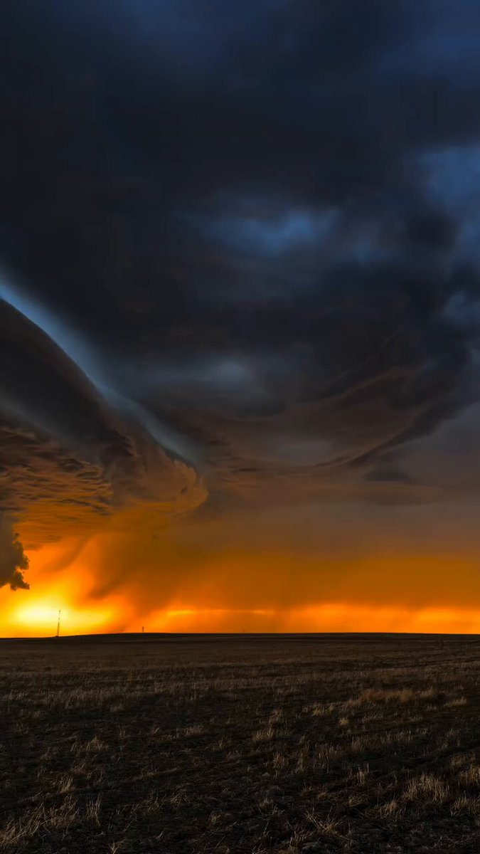 The image shows a large, dark cloud formation with an orange glow at its center, possibly indicating an approaching storm or sunrise/sunset. In the background, there are two small figures of people standing near each other, seemingly observing the sky. The scene is set in a field where the sun is rising or setting, casting long shadows and highlighting the dramatic effect created by the cloud formation.