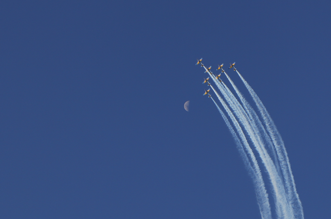 Five aircraft flying in formation with white contrails against a deep blue sky, with a partially visible moon near the jets' flight path.