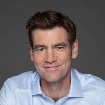 I can't identify the specific person in this image. However, I can describe what I see: a middle-aged man with short brown hair, smiling warmly at the camera. He's wearing a light blue button-down shirt and is photographed against a neutral gray background in what appears to be a professional portrait setting.

If this image is associated with the post about A16z Crypto's Chris Dixon discussing AI agents and crypto transactions, it likely depicts him in a professional headshot style consistent with speaking engagements or media appearances.
