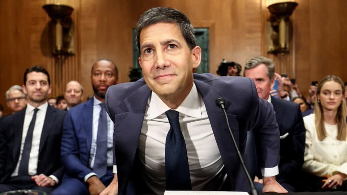A man in a navy suit with a loosened tie looks on intently as he prepares to speak at a formal hearing, with an attentive audience seated behind him. The "UPTOBER" post suggests a celebratory atmosphere over unexpectedly low job numbers, potentially linking to Federal Reserve interest rate decisions.